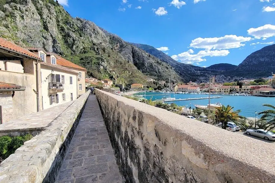 a walkway on top of Kotor city walls with a stunning view of Kotor bay & old houses with â›° backdrop on the other side